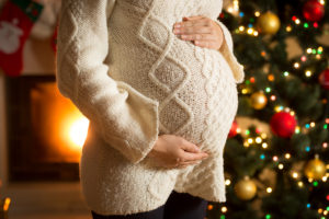 A pregnant woman poses for a photo in front of a Christmas tree.