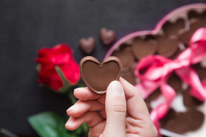 A person holds a heart-shaped piece of chocolate. A rose and more chocolate are shown in the background.