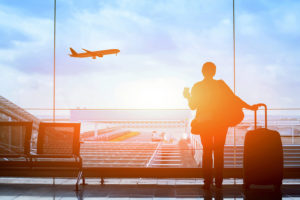 A person stands at an airport and looks out the window to watch a plane fly in the air. The person holds a suitcase.