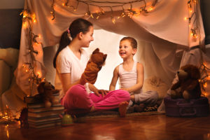 A young girl and boy sit inside an indoor fort made of blankets. They smile big as they play with puppets. 