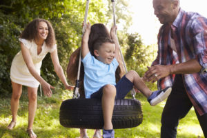 A kid sits on a tire swing as three people push the swing.