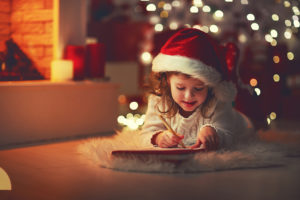 A young girl wears a Santa hat and appears to be writing a list in front of a Christmas tree.