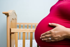 A pregnant woman stands next to a crib and holds her belly.