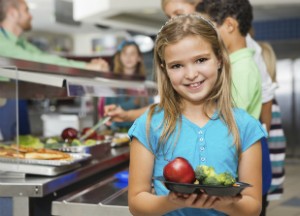 A young girl grabs her school lunch that contains an apple and broccoli. She poses for a photo and smiles in a lunchroom.