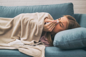 A woman lies on a blue couch and covers herself with a blanket. She blows her nose with a tissue.