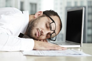 A man falls asleep at his work desk with his laptop wide open. He appears to have work sleep issues.