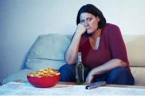 A woman sits on a couch and watches TV. A table in front of her holds a remote, a bottle of beer and a bowl full of chips.