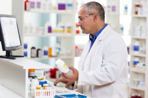 A pharmacist looks at a pill bottle behind the counter of a pharmacy.