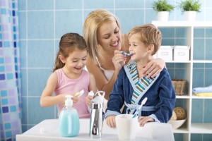 A mother helps her son and daughter brush their teeth in a bathroom.