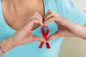 A woman shapes her hands into a heart. The woman is wearing a red ribbon that symbolizes heart disease.