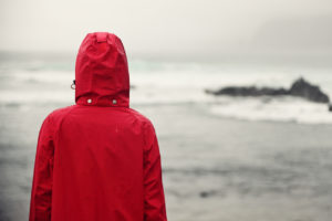 A woman wearing a red raincoat stares into the ocean on a gloomy day.