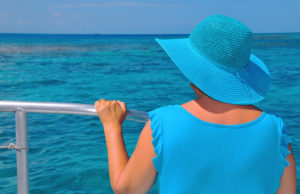 A woman wearing a blue shirt and blue sun hat holds a railing of a boat and stares into the ocean.
