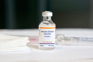 A small glass vaccine vial with a shingles vaccine label is shown on a table along with a syringe. 