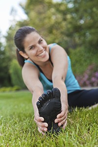 A woman stretches out her legs as she sits in grass. She wears barefoot running shoes.