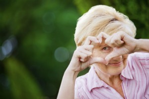 An elderly woman poses for a photo and shapes her hands to create a heart over her eye.