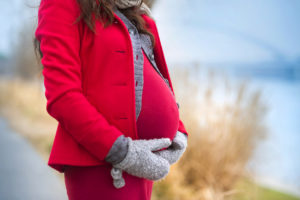 A pregnant woman holds her belly as she stands outside. She wears a red winter coat and gray gloves.