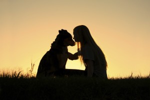 A young girl hugs her dog outside as the sun sets in the background.