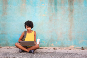 A young woman sits on the ground and works on her laptop.