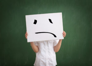 A child holds up a sign to cover their face. The sign shows a frowning face.