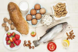 Bread, nuts, eggs, fish, strawberries, a lemon, a glass of milk and a tomato is shown.