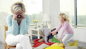 A mother holds her head in her hands as she appears to be having a migraine. Her children are playing with pillows in the backgrond.