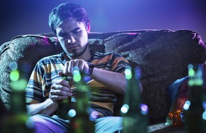 A man sits on a couch and opens a bottle of beer. He appears upset.