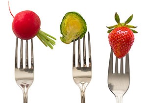 Three forks are shown, each hold a different food item. One fork holds a radish, another fork holds a brussel sprout, and the last fork holds a strawberry.