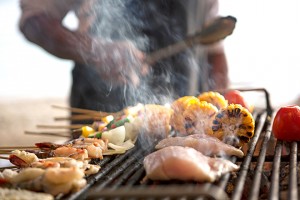 A man cooks corn, chicken and kabobs on a grill.