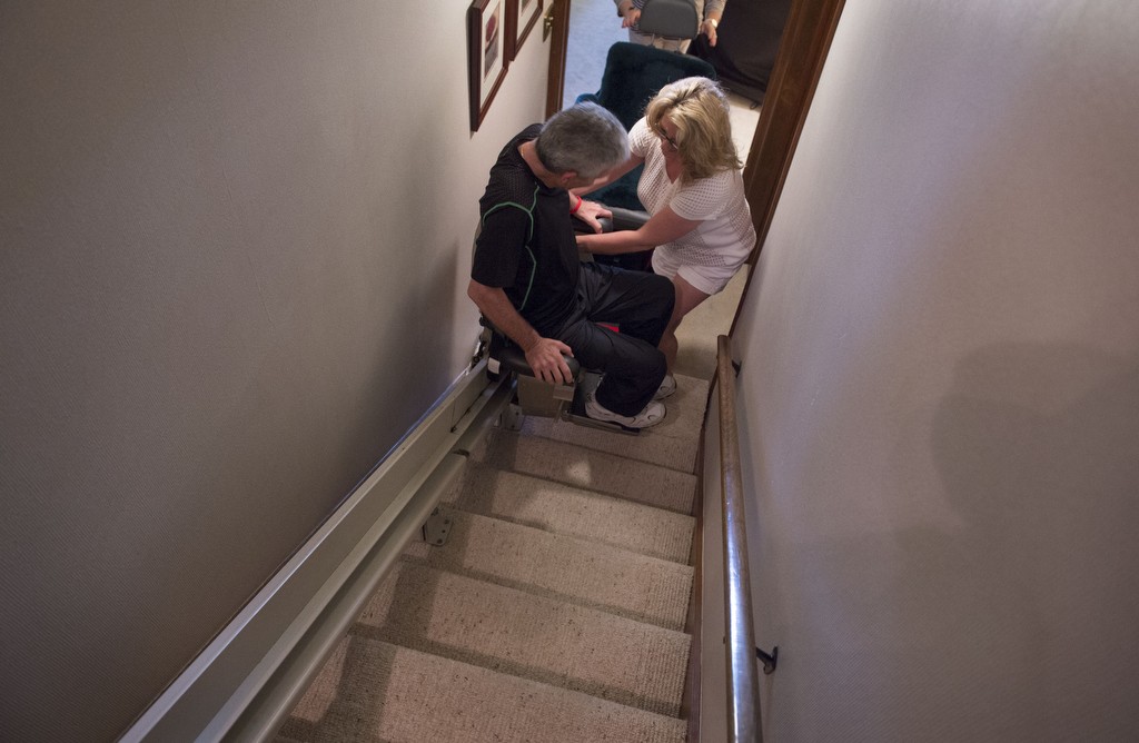 Todd Alward gets in the stair chair that has been installed in his mother's home. His sister, Leisha McCullough, helps him get buckled in in order to go up a flight of stairs.