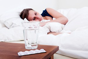 A woman lies in bed and appears awake. Her bedside table holds a glass of water and medication.