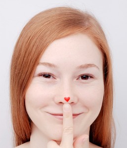 A woman smiles as she places her hand on her nose. Her nose has a red, heart sticker on it.