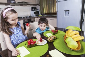 A young boy and girl eat strawberries in a kitchen.