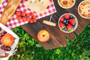 A picnic table holds a baguette, cheese and fruit.