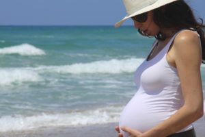 A pregant woman stands near the beach wearing a hat and sunglasses.