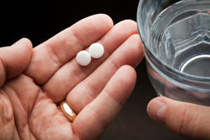 A person holds two aspirin tablets in one hand, and a glass of water in the other.