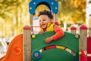 A young boy smiles big as he plays in an outdoor playground.
