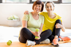A woman wraps her arms around another woman. They sit on a yoga mat with dumbbell weights and smile.