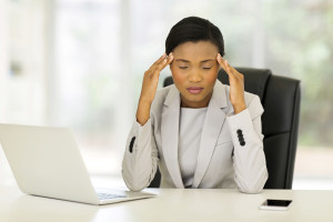 A woman holds her hands to her forehead at work. She sits at a desk with a computer.