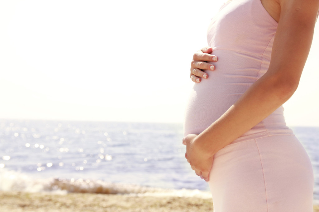 A pregnant woman holds her belly as she stands near the beach.