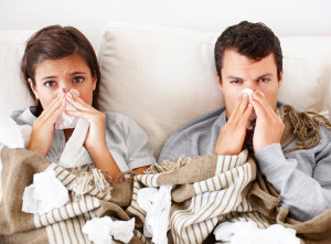 A woman and man lie in bed with tissues covering their bed. They both are blowing their nose.