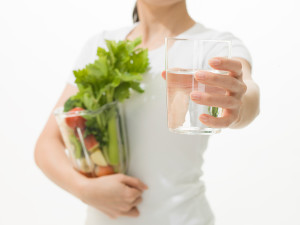 A woman holds a glass of water and holds it out in front of her. She also holds a blender with vegetables in her other arm.