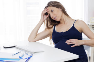 A pregnant woman leans her arm on a table and holds her belly. She appears uncomfortable.