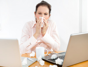 A woman appears sick as she works. The woman blows her nose into a tissue. Multiple tissues lie next to her laptop.