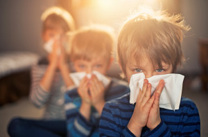 Three young boys blow their nose with a tissue.