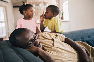 A man appears asleep on his couch as two children tell each other to be quiet.