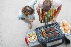 A man cooks chicken and burgers on a grill as a woman looks on.
