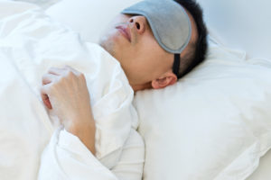 A man lies in bed and covers his eyes with an eye mask.