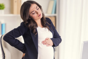 A pregnant woman is standing and holds her belly and appears uncomfortable.