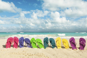 Six pairs of colorful flip-flops sit in the sand near a beach.