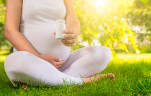 A pregnant woman sits in the grass outside and holds a pill bottle.